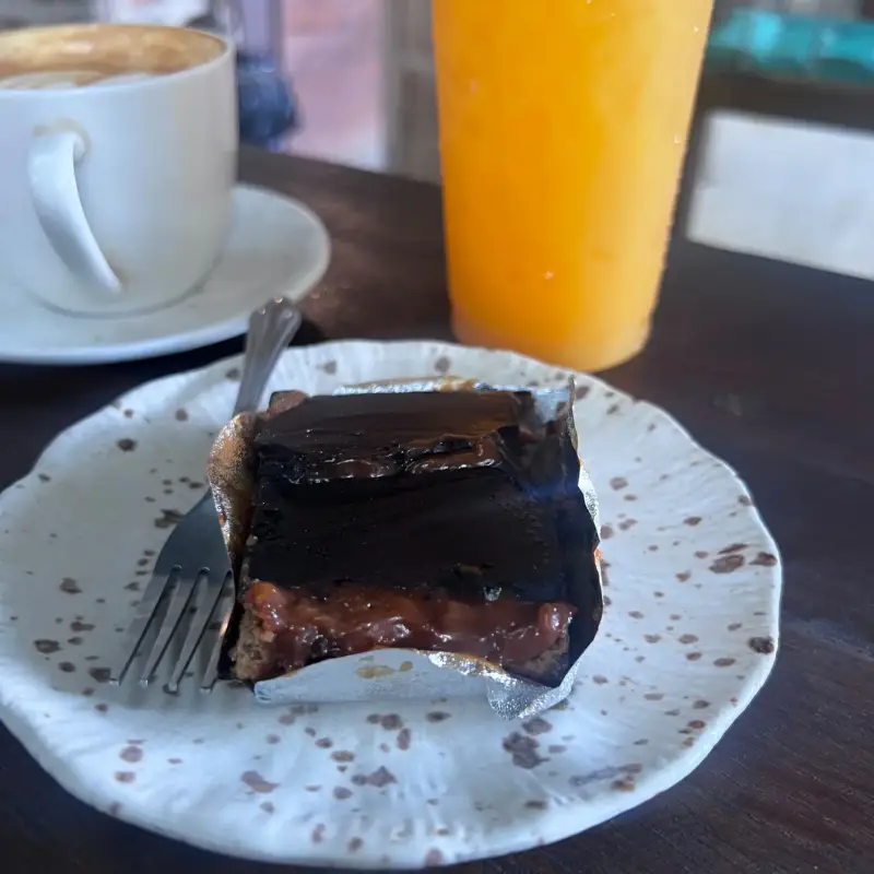 Kuchenstück mit dunkler Glasur neben Kaffee und einem orangefarbenen Getränk