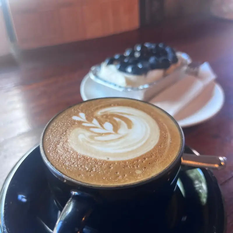 Kaffee mit Latte Art in einer dunklen Tasse, dahinter ein Beerendessert